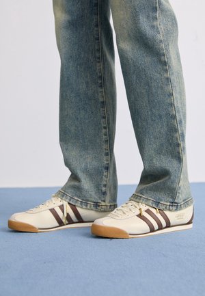 Personne portant un jean bleu et des baskets blanches Adidas avec des bandes marron, debout sur un sol bleu clair sur fond blanc.