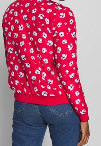 Veste bomber rouge à motif floral avec une finition satinée, poignets et taille côtelés ; ornée de petites fleurs bleues et blanches.