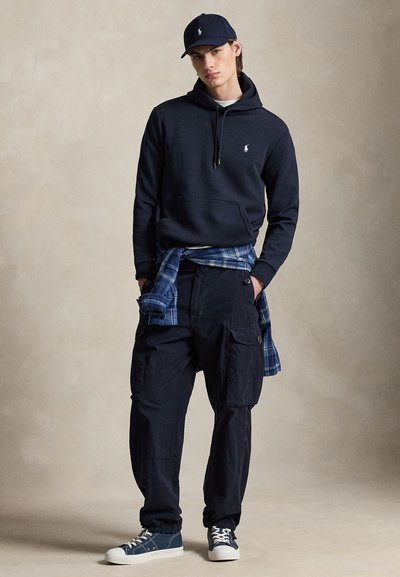 Jonge man die een marineblauwe hoodie draagt, bijpassende pet, cargobroek, sneakers en een blauw geruit overhemd dat om de taille is gestrikt, staand tegen een neutrale achtergrond.