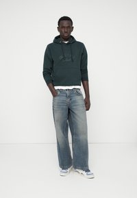 Hoddie vert foncé avec lettrage en relief, superposé sur une chemise blanche. Jeans en denim bleu à jambes larges avec une finition délavée et des baskets blanches.