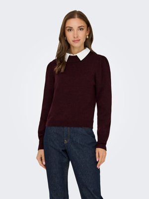 Kvinde iført en mørk vinrød sweater med en hvid skjorte med krave under og mørkeblå jeans, stående mod en ensfarvet lys baggrund.