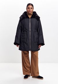 Sort pufferjakke med en kunstlæderkrave, lynlås og to forlommer. Styllet med brede beige bukser og sorte slip-on sko.