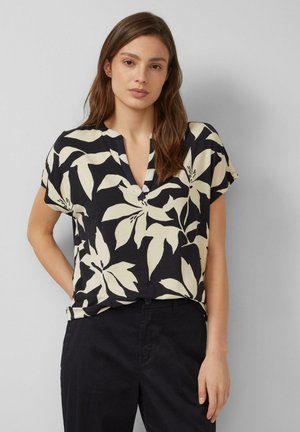 Kobieta o długich brązowych włosach, ubrana w czarno-beżową bluzkę w kwiaty z krótkim rękawem oraz czarne spodnie, stojąca na tle jednolitego szarego tła.