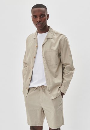 Camicia beige con bottoni e tasca, indossata sopra una maglietta bianca, abbinata a pantaloni corti beige con coulisse. Tessuto morbido, design casual.
