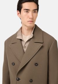 Abrigo de lana marrón de doble pechera con grandes solapas, botones negros y un cuello texturizado superpuesto a una camisa beige.