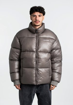 Chaqueta tipo abrigo en marrón con cuello alto, secciones acolchadas y puños de punto en gris. Presenta una cremallera frontal y un logo en el pecho.