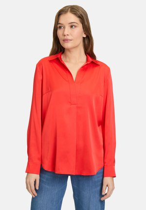 Blouse en satin rouge avec col, manches longues et design à patte de boutonnage. Texture lisse et coupe ample, associée à un pantalon en denim bleu.
