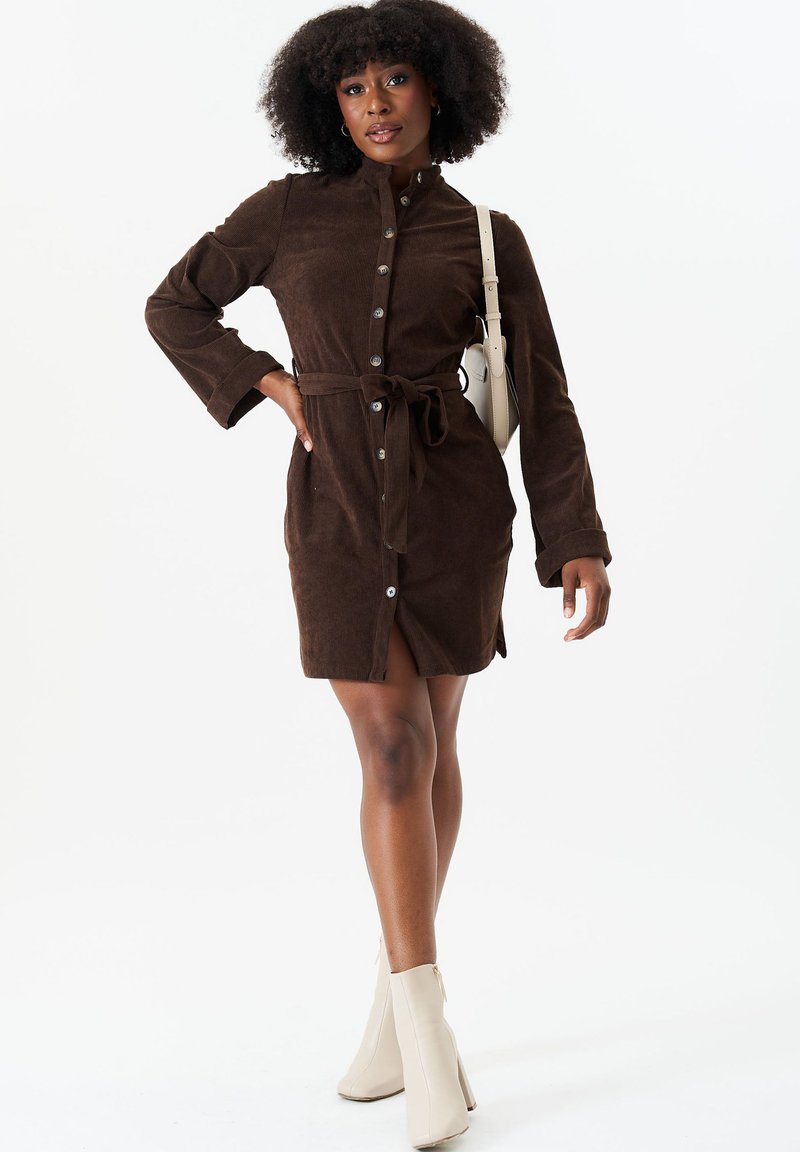 Brown corduroy shirt dress with long sleeves, button front, and tie waist. Features side slit and paired with beige ankle boots.