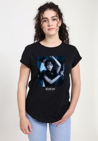 Žena s kudrnatými vlasy, která má na sobě černé tričko s obrázkem tmavovlasé dívky tančící a nápisem "Středa" pod obrazem.