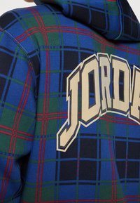 Sudadera con capucha que presenta un diseño de cuadros en azul, verde y rojo. Logo blanco "JORDAN" exhibido prominentemente en la parte posterior. Textura suave.