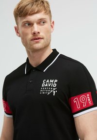 Polo nero con colletto bianco. Presenta maniche rosse con il numero 19 e branding. Il materiale appare texturizzato e traspirante.