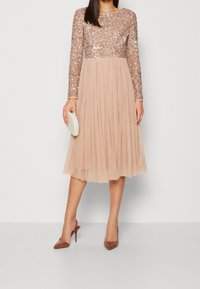 Abito a maniche lunghe con top a paillettes in oro rosa e gonna plissettata in tulle beige, abbinato a una clutch bianca e tacchi marroni.