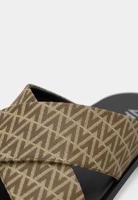 Flat sandal med en brunmønstret stropp og svart såle. Stroppen har et geometrisk design i en lysere nyanse. Enkel og funksjonell.