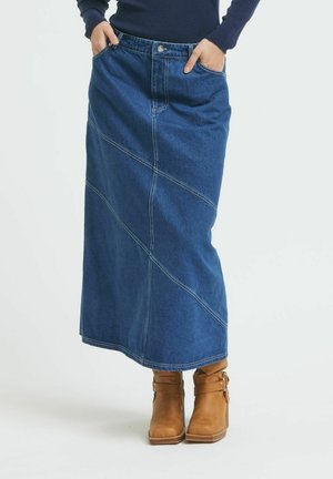 Maxi-Denimrock in Dunkelblau mit diagonaler Naht, ausgestattet mit vorderen Taschen und einem Knopfverschluss. Tragen mit braunen Stiefeletten.
