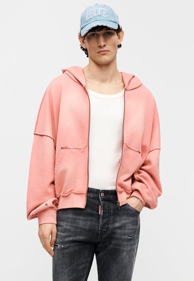 Ροζ φούτερ με φερμουάρ και άνετη εφαρμογή, δύο μπροστινές τσέπες και ακατέργαστες ραφές, συνδυασμένο με λευκό πουκάμισο και σκισμένο σκούρο τζιν.