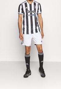 Maglia a righe bianche e nere della Juventus con maniche corte, abbinata a pantaloni corti bianchi e calze nere con tre righe bianche. Scarpini da calcio neri.