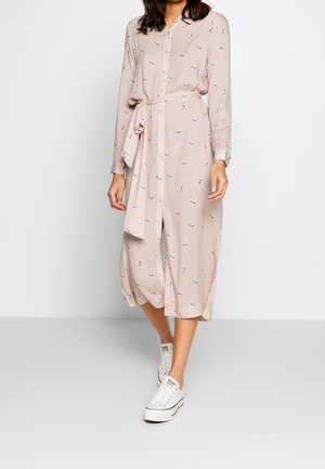 Robe longue boutonnée de couleur rose pâle avec motif floral, ceinturée à la taille, associée à des baskets blanches pour un look décontracté.
