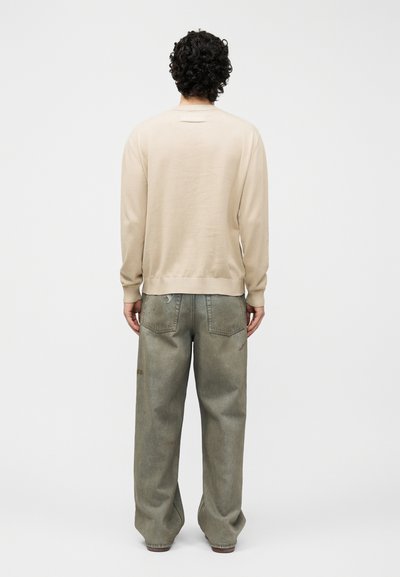 Άνδρας με σγουρά σκούρα μαλλιά στέκεται με την πλάτη, φορώντας μπεζ μακρυμάνικο πουκάμισο και χαλαρά ξεθωριασμένα πράσινα τζιν με πίσω τσέπες.