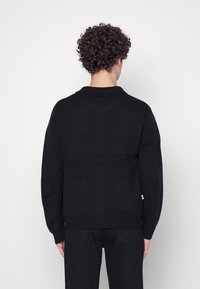 Svart stickad tröja med en mjuk textur, som har en sjalkrage och en avslappnad passform. Ribbstickade ärmar och fåll med en accenttag på sidan.