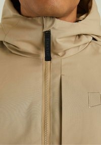 Beżowa kurtka z kapturem, z wysokim kołnierzem, centralnym zamkiem błyskawicznym i czarną metką z logo. Gładki materiał o delikatnym połysku z minimalną ilością szwów.