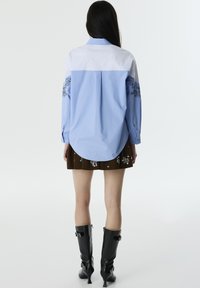 Camicia azzurra con bottoni, colletto a righe e ricami floreali sulle maniche, abbinata a una gonna plissettata marrone e stivali al ginocchio neri.