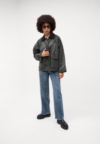 Kaklo didelio dydžio švarkas su dviem priekinėmis kišenėmis, derinamas su šviesiai mėlynomis tiesiomis kelnėmis ir juodais platforminiais batais, ant balto fono.