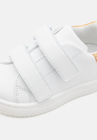 Chaussure en synthétique blanche avec deux brides Velcro, bout arrondi, texture lisse et un accent jaune contrastant au talon. Semelle en caoutchouc.