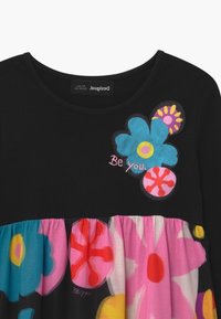 Czarna sukienka z kolorowymi kwiatowymi naszywkami i haftem z napisem "Bądź sobą" na klatce piersiowej, z marszczonym dołem w wyrazistych wzorach kwiatowych.