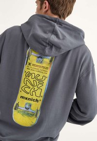 Felpa grigia con un grafico di skateboard sul retro, evidenziato in giallo con testo e dettagli neri. Tessuto morbido, vestibilità rilassata.