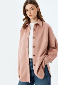Rosa fuskmocka skjortjacka med krage, framknäppning, två fickor och lång ärm, i kombination med en vit topp och blå jeans.