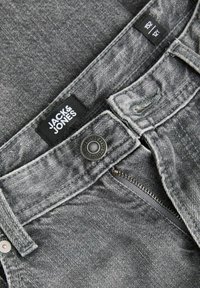 Gros plan sur la ceinture d'un jean denim gris montrant l'étiquette Jack & Jones, un bouton en métal, une fermeture éclair et une étiquette de taille 152 12Y.