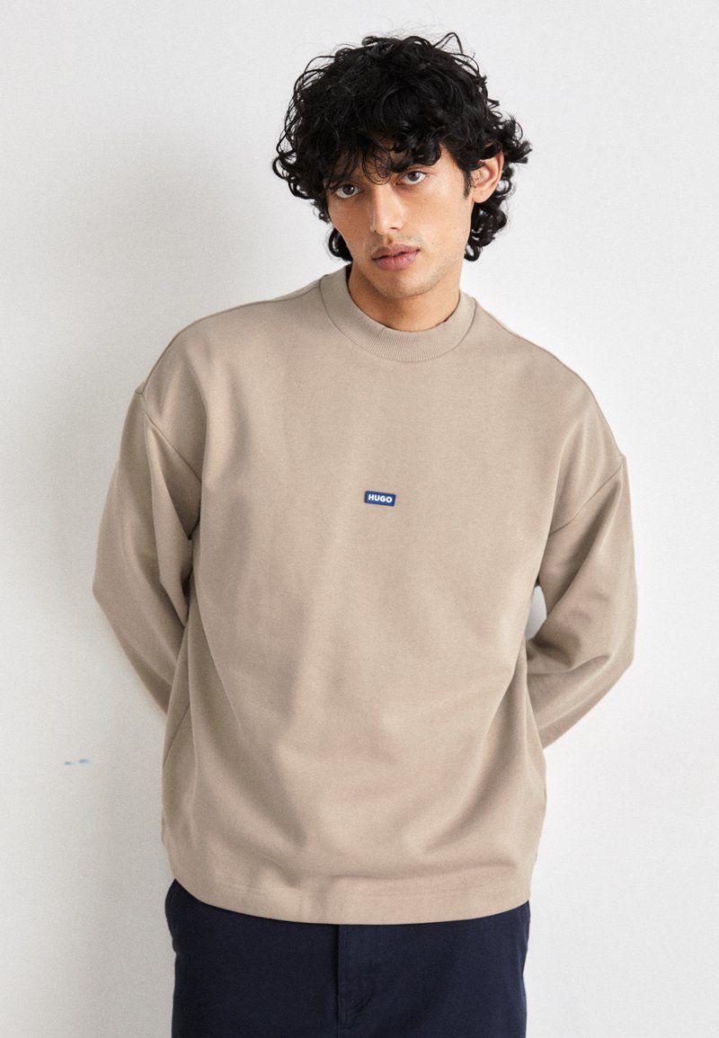 Felpa beige con un collo alto, spalle cadenti, polsini a coste e un piccolo logo blu HUGO sul petto. Tessuto morbido e textureata.