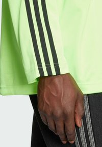 Neona zaļa garu piedurkņu krekls ar melnu trīs strīpu dizainu uz piedurknes; izgatavots no teksturētas auduma un ar brīvu piegriezumu.