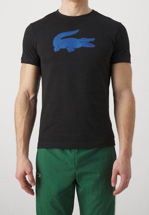 Homme portant un t-shirt noir à manches courtes avec un grand motif de crocodile bleu et un pantalon vert avec un petit logo crocodile noir et blanc.