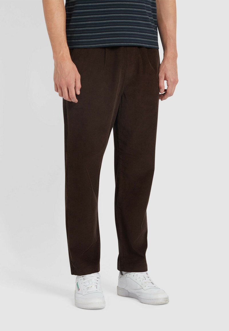 Pantaloni in velluto a coste marroni con una vestibilità rilassata, caratterizzati da pieghe frontali e una superficie testurizzata. Abbinati a sneakers bianche per creare un contrasto.