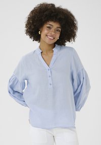 Camicia button-up azzurra chiara con colletto arricciato e maniche lunghe a sbuffo, realizzata in un morbido tessuto testurizzato. Presenta tre bottoni.