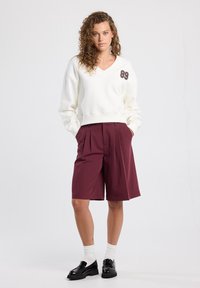 Kerman v-pääntie sisäpuolella, jossa on viininpunainen numero yksityiskohta, yhdistetty viininpunaisiin pleatattuihin shortseihin. Mustat loafers-kengät ja valkoiset sukat täydentävät asua.