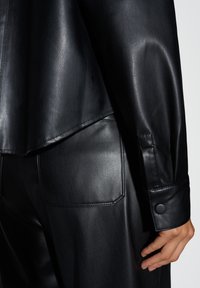 Giacca di pelle nera con texture liscia, colletto appuntito e polsini con bottoni. Il design include un orlo posteriore arrotondato e cuciture strutturate.