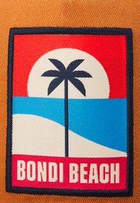 Gesticktes Abzeichen mit einer Palme, rotem und blauem Hintergrund und Sand. Der Text lautet "BONDI BEACH" in fetter, cremefarbener Schrift.