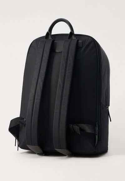 Mochila de nylon negra con correas de hombro acolchadas, mango en la parte superior y cierre de cremallera. Textura suave y diseño minimalista. Detalle en el bolsillo lateral.