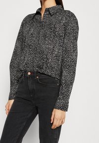 Musta napitettava paita, jossa on valkoinen pilkullinen kuvio, pitkät hihat ja klassinen kaulus. Yhdistetty korkeavyötäröisiin mustiin farkkuihin.