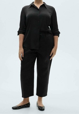 Persona que lleva una camisa negra holgada con botones y mangas remangadas, pantalones negros de pierna recta y zapatos planos negros con detalles de ojales.