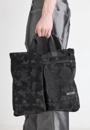 Personne tenant un sac cabas noir à motif camouflage avec des poignées courtes, portant un pantalon gris foncé sur un fond clair.
