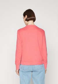 Sudadera color coral con mangas largas, puños y dobladillo acanalados. Textura suave. El diseño en la parte posterior presenta un pequeño punto naranja cerca del escote.