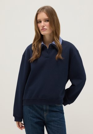 Giovane donna con lunghi capelli castani che indossa un maglione blu navy con colletto a quadri blu e jeans blu, in piedi davanti a uno sfondo semplice.