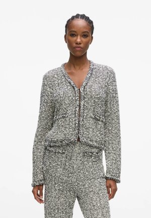 Kvinde iført en grå og hvid struktureret strikket jakke og matchende bukser, stående med et neutralt udtryk mod en ensfarvet hvid baggrund.