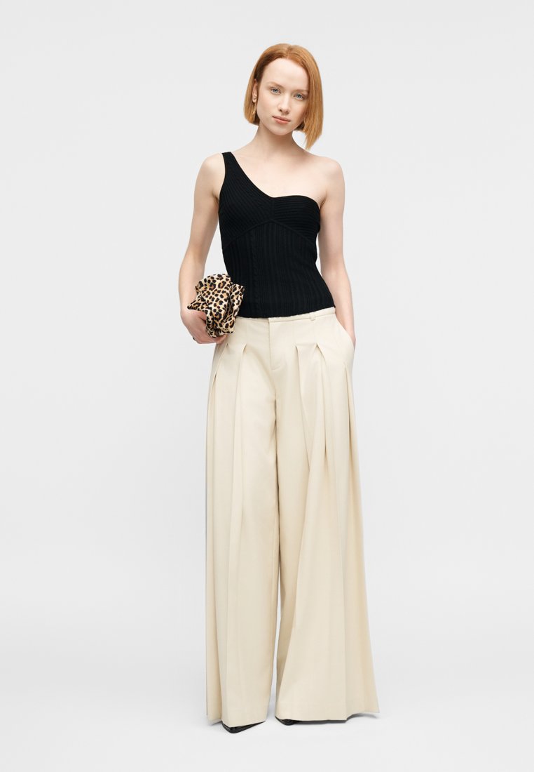 Haut noir côtelé à une épaule associé à un pantalon large beige avec plis à l'avant. Tient une pochette à imprimé léopard.