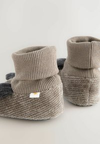 Gestrickte beige Hausschuhe mit einem gerippten Bund, grauen Akzenten, strukturierter Oberfläche und einem weißen Etikett mit einem Sonnenlogo.