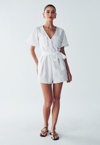 Baltas trumpas jumpsuit'as su V formos iškirpte, saga priekyje, dviem krūtinės kišenėmis ir diržu juosmens srityje. Dėvimi su juodomis plokščiomis sandalais.