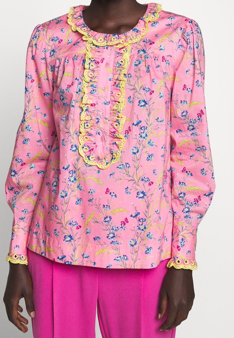 Rosa blus med blommönster och gul spetsdetalj, med långa ärmar, en volang framtill och en avslappnad passform, kombinerad med livfulla rosa byxor.
